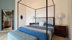 Ett sovrum med en himmelssäng, ett nattduksbord med en lampa, ett litet bord vid sängen med en lampa, en dekorativ detalj som hänger på väggen och en krukväxt.