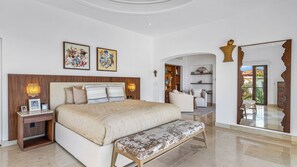A spacious bedroom with a large bed, bedside tables, and a bench. There are two framed artworks on the wall, a wooden headboard, and a large mirror with a decorative frame. - Stunning 5BR Oceanview Villa in Conchas Chinas (Puerto Vallarta)