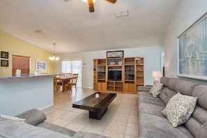 A spacious living room with a sofa, coffee table, and TV stand. There is a dining area with a table and chairs. The room has a ceiling fan and a large window. - Best Value Near Disney! Private Pool, BBQ, Fenced Yard & Room for 8 (Kissimmee)