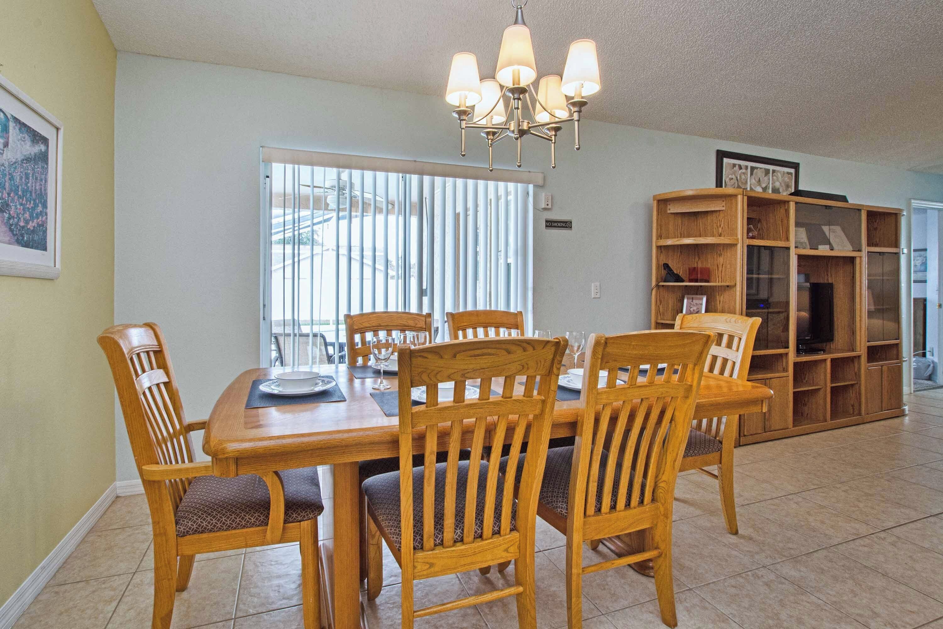 A dining area with a wooden table and chairs, a chandelier, a bookshelf, and a window with blinds.