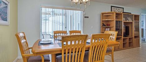 A dining area with a wooden table and chairs, a chandelier, a bookshelf, and a window with blinds.