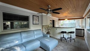 A living room with a blue leather sofa, a ceiling fan, and a kitchen area with two bar stools. - Charming 1-bedroom Guesthouse in Amazing Ozona Free Parking, Walk to Restaurants (Palm Harbor)