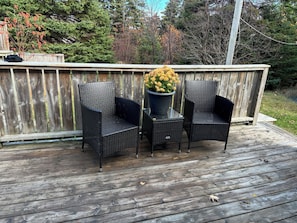 Een houten terras met twee rieten stoelen en een tafel, omgeven door een houten omheining en bomen.