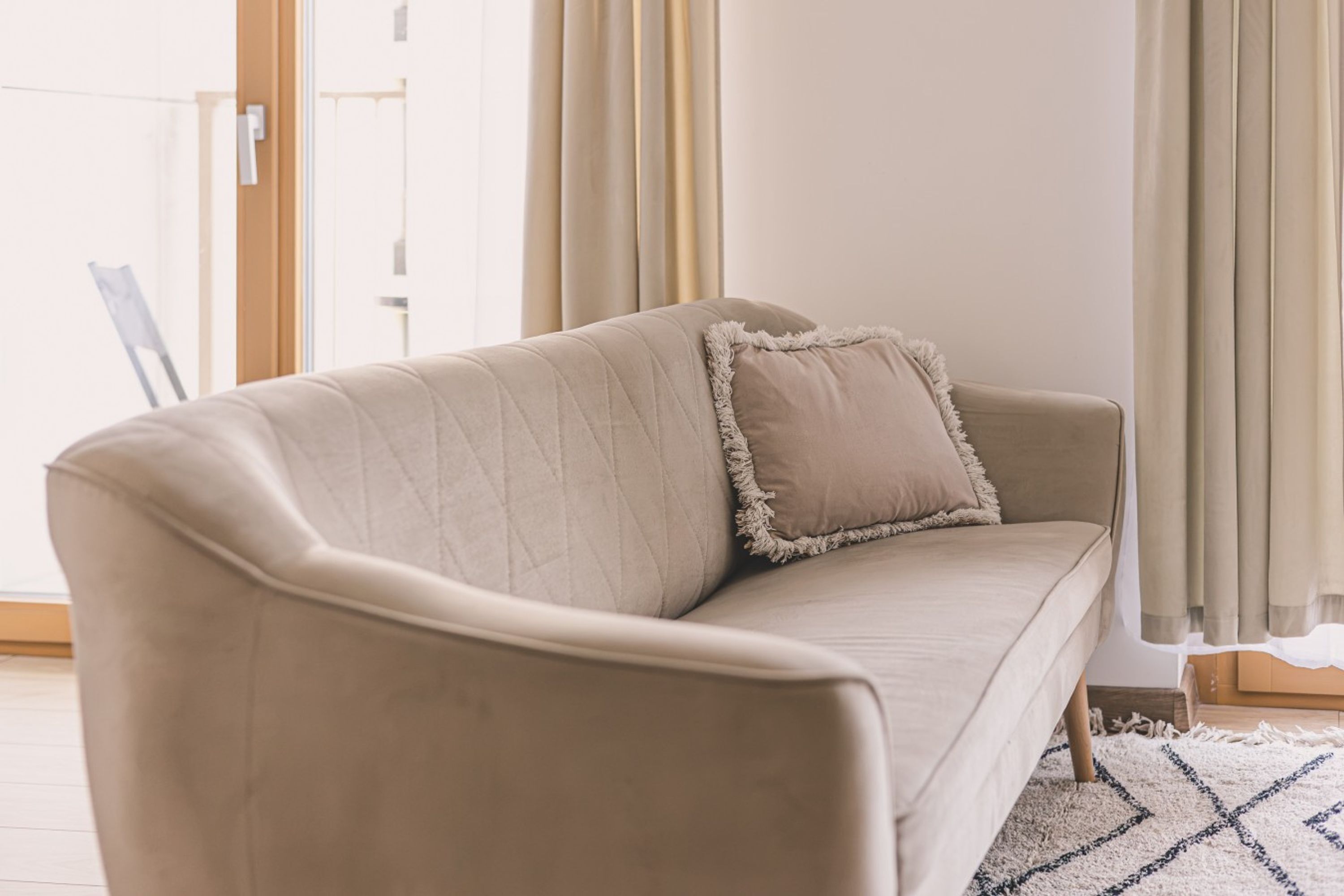 Un sofá color beige con respaldo acolchado y un solo cojín, ubicado en una habitación con un ventanal y cortinas.