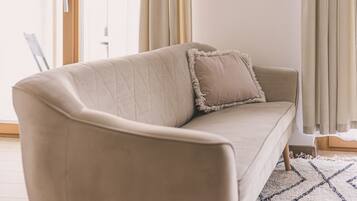 Un sofá color beige con respaldo acolchado y un solo cojín, ubicado en una habitación con un ventanal y cortinas.