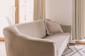 Un sofá color beige con respaldo acolchado y un solo cojín, ubicado en una habitación con un ventanal y cortinas.