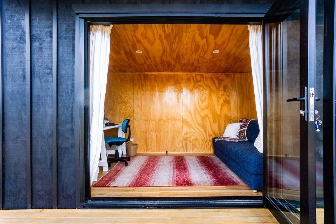 A room with a wooden ceiling, a bed with a blue cover, a desk with a chair, and a red and white striped rug.