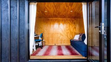 A room with a wooden ceiling, a bed with a blue cover, a desk with a chair, and a red and white striped rug.
