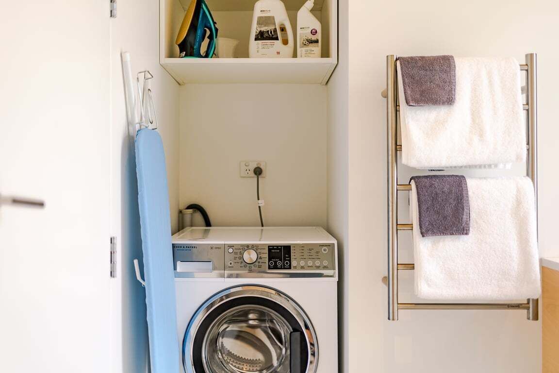 A compact laundry area with a washing machine, a drying rack, and a shelf with laundry products.