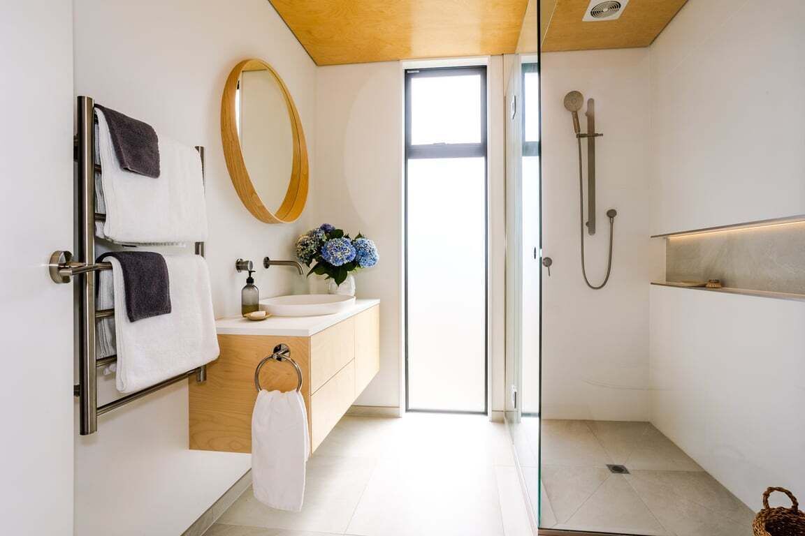 A modern bathroom with a glass shower enclosure, a wooden vanity, a round mirror, and a view of the outdoors.