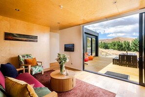 A modern living room with a wooden ceiling, a flat-screen TV mounted on the wall, a round coffee table with a vase of flowers, and a large sliding glass door leading to an outdoor deck.