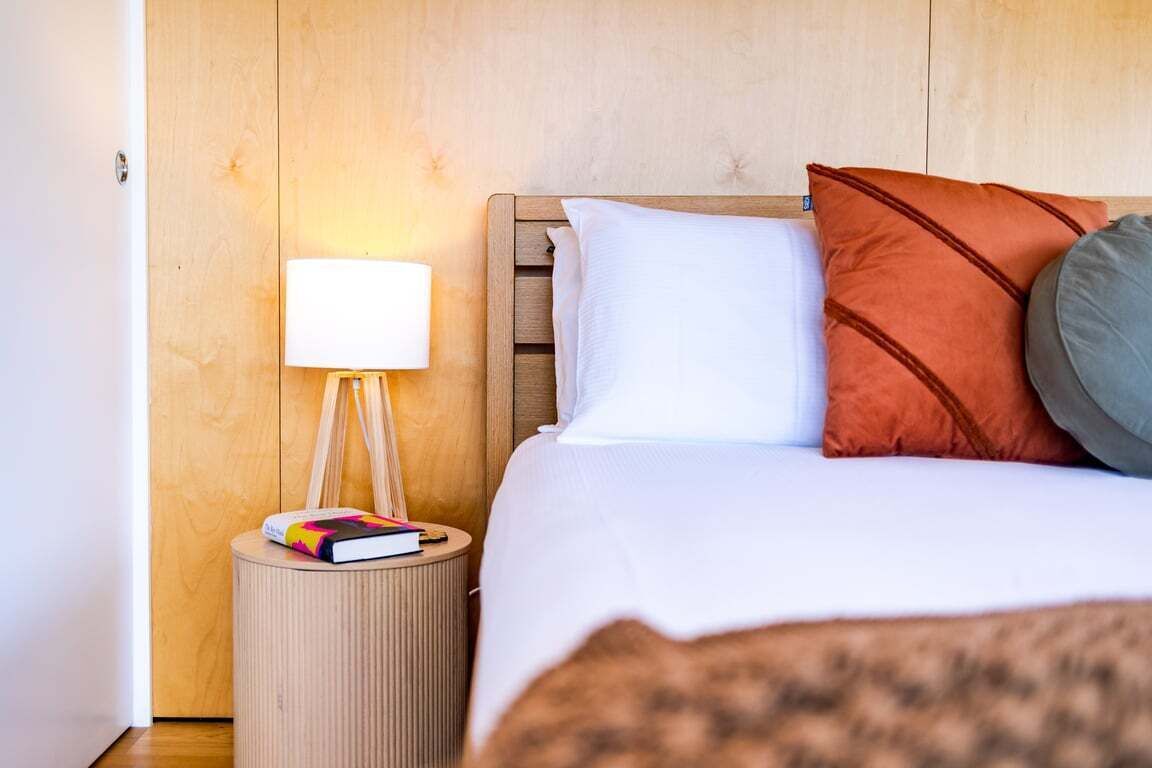 Een keurig opgemaakt bed met witte en oranje kussens, een bedlampje en een boek op een rond nachtkastje.