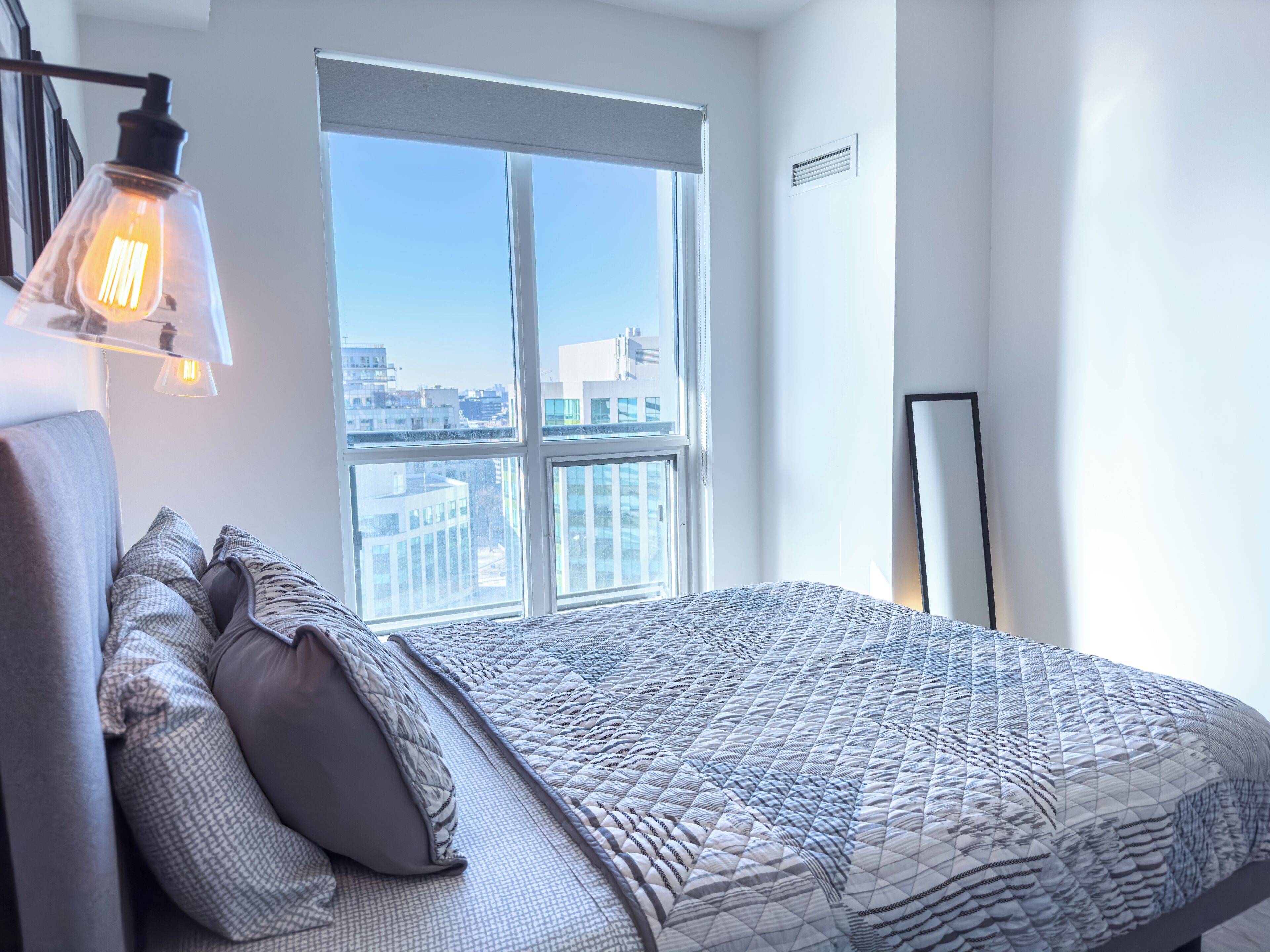 A modern bedroom with a large window, a bed with patterned bedding, and a wall-mounted light fixture.