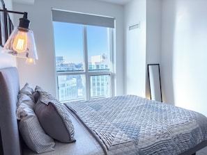 A modern bedroom with a large window, a bed with patterned bedding, and a wall-mounted light fixture. - Central Suites Downtown Toronto (Toronto)