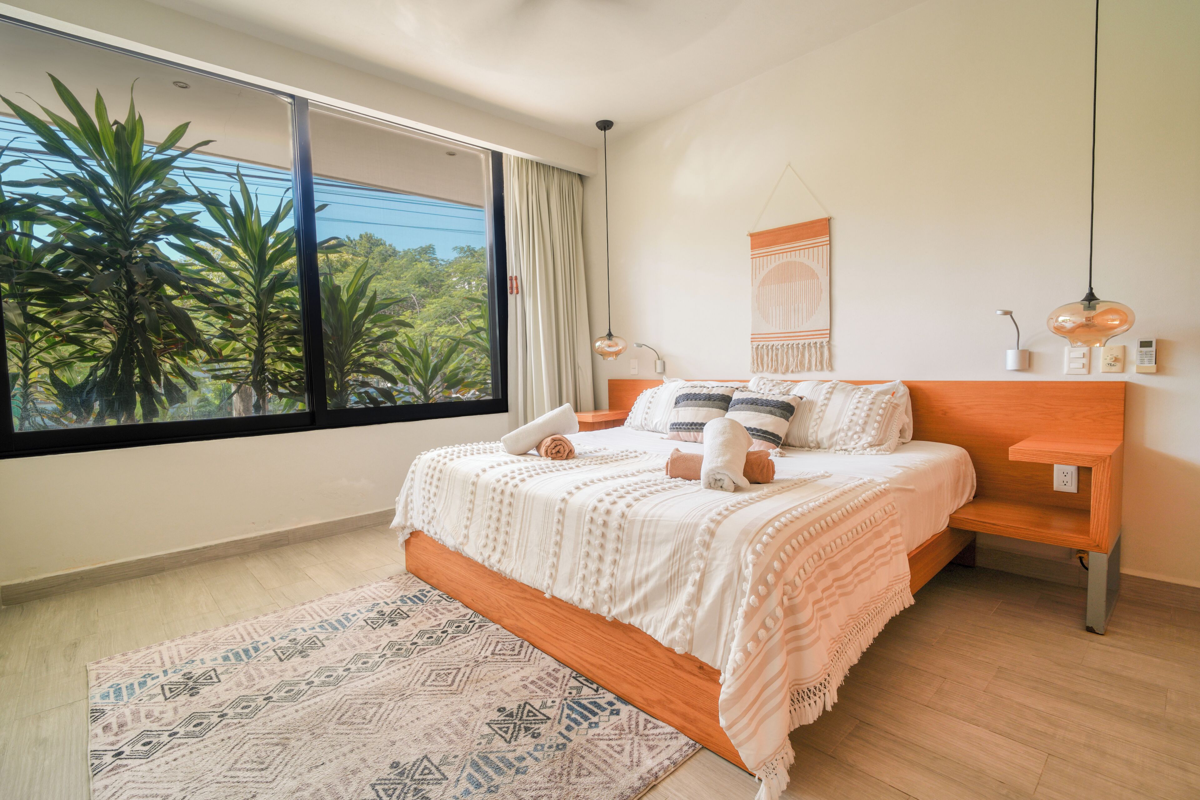 A bedroom with a large window, a bed with white bedding, a wooden nightstand, and a wall-mounted lamp.