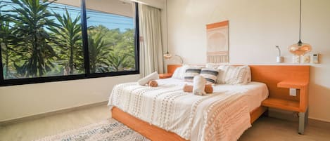 A bedroom with a large window, a bed with white bedding, a wooden nightstand, and a wall-mounted lamp.
