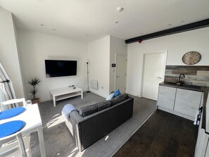 Ein modernes Wohnzimmer mit einem grauen Sofa, einem Flachbildfernseher, einem weißen Couchtisch und einer kleinen Pflanze.