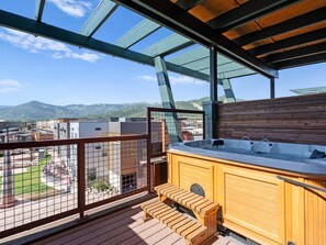 Eine Holzterrasse mit Whirlpool und Sitzgelegenheit, mit Blick auf eine gebirgige Landschaft.