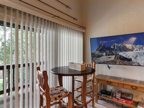 A room with a round table, two chairs, a bookshelf, and a large window with blinds. - Snow Flower 124 | Ski-In/Ski-Out | Loft | Pools & Hot Tub (Park City)