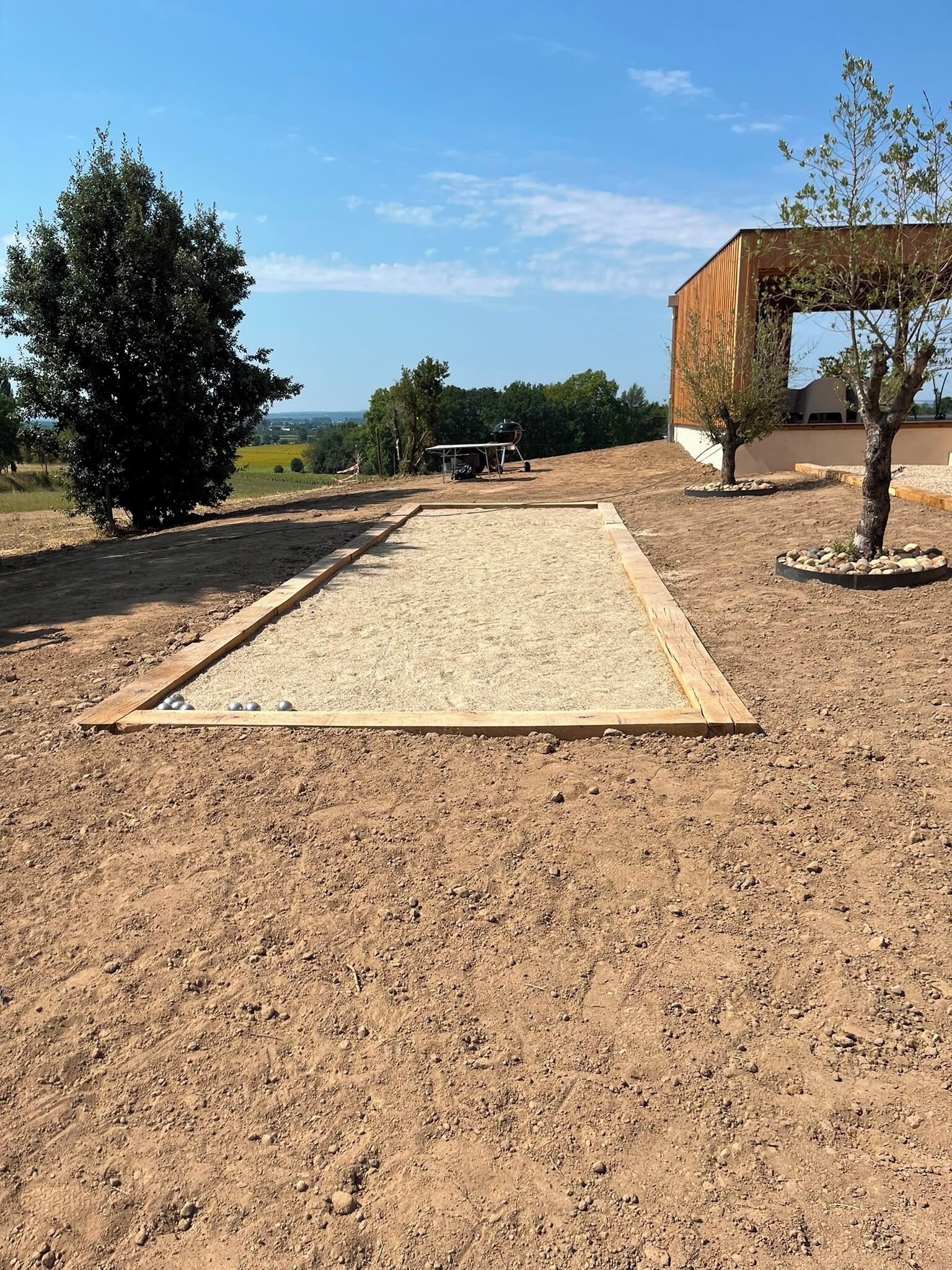 Un espace extérieur recouvert de terre, avec une structure en bois, des arbres et un bâtiment en arrière-plan.