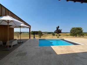 Ein Schwimmbecken mit Holzterrasse und Sitzgelegenheiten im Freien, umgeben von blauem Himmel und einer weiten Landschaft.