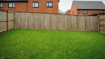A backyard with a wooden fence, a grassy lawn, and two brick houses with pitched roofs.