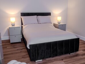 Ein ordentlich bezogenes Bett mit schwarzem Kopfteil und weißer Bettwäsche, begleitet von zwei Nachttischen mit Lampen, in einem Zimmer mit Holzboden.