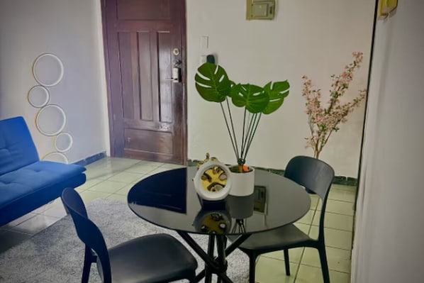A room with a round table, two chairs, a blue sofa, a wooden door, and a potted plant with large leaves.