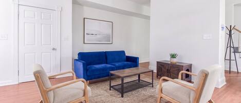 Un salon avec un canapé bleu, deux fauteuils, une table basse, un tapis, un meuble orné d’une plante et un ventilateur de plafond.