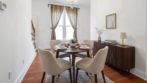 Uma sala de jantar com uma mesa redonda posta para quatro pessoas, um lustre, um aparador com livros e um espelho na parede.