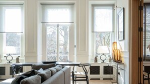 A living room with a sofa, a table, and two lamps. There are large windows with a view of trees. - Cadogan Square 2-bed Direct Elevator Access (London)