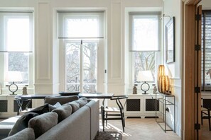 Ein Wohnzimmer mit einer Couch, einem Tisch und zwei Lampen. Große Fenster bieten Blick auf Bäume.