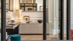 A modern interior with a glass door leading to a hallway, a desk with a lamp, and a blue ottoman. - Cadogan Square 2-bed Direct Elevator Access (London)