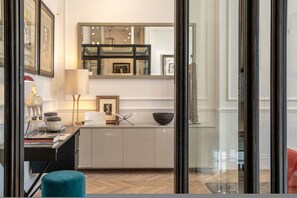 Ein modernes Interieur mit einer Glastür, die zu einem Flur führt, einem Schreibtisch mit Lampe und einem blauen Pouf.