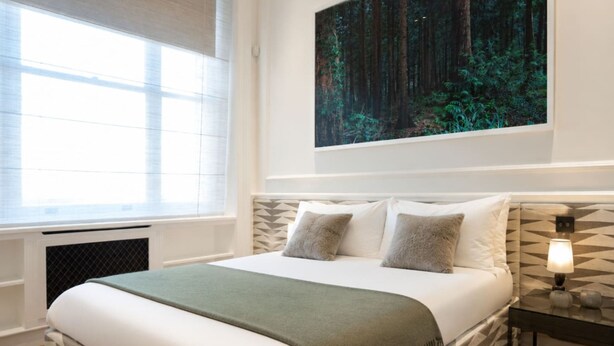 A neatly made bed with a green throw, a large window with blinds, and a forest-themed painting on the wall. - Cadogan Square 2-bed Direct Elevator Access (London)