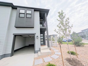 A two-story residential building with a garage, a balcony, and a landscaped front yard with stepping stones and small trees. - Modern 1BR/1BA with Attached Garage private entrance and Patio (Lone Tree)