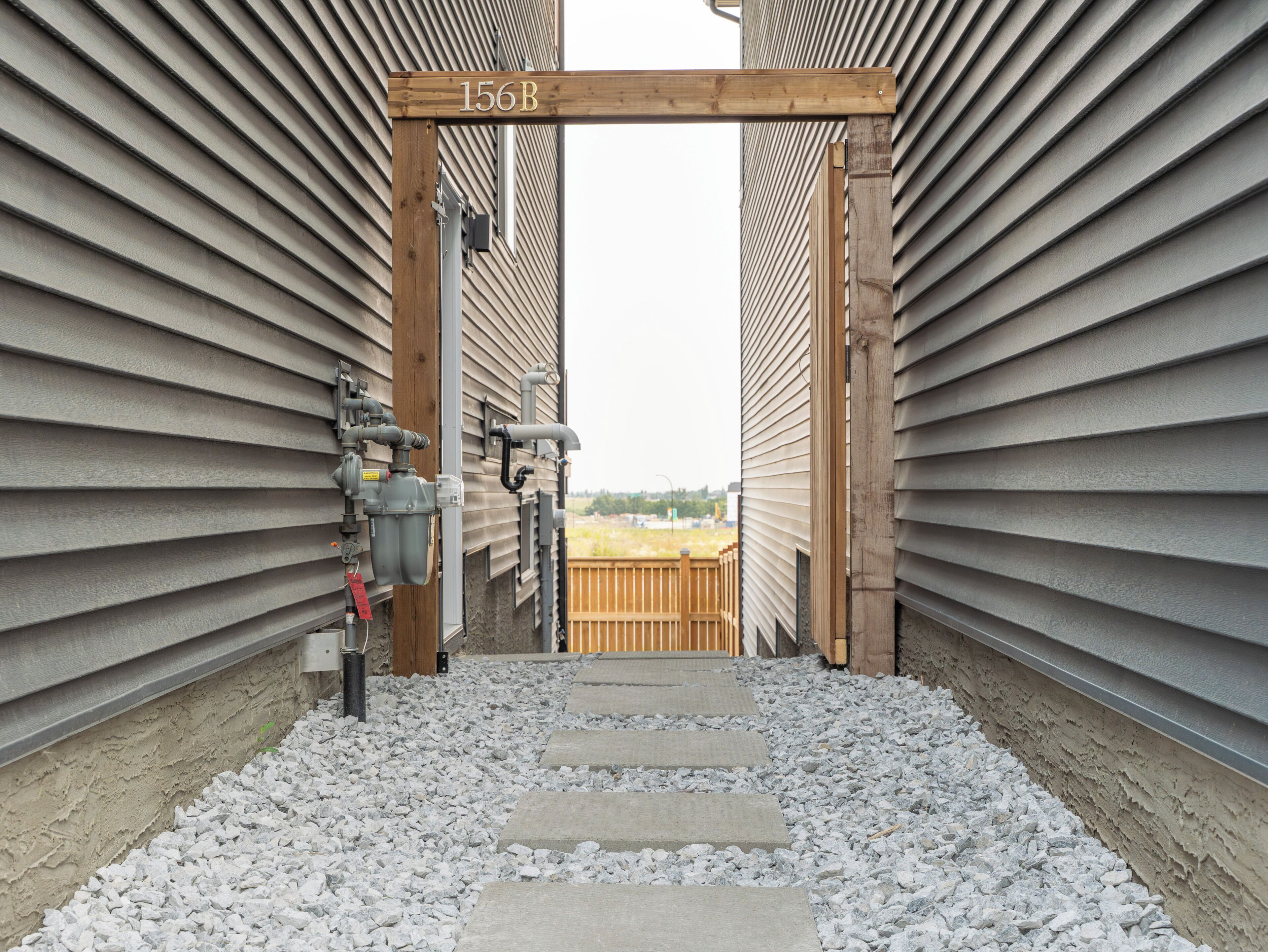 A pathway leading to a wooden archway with the number 156B, flanked by siding walls, with outdoor plumbing and a wooden deck in the background.