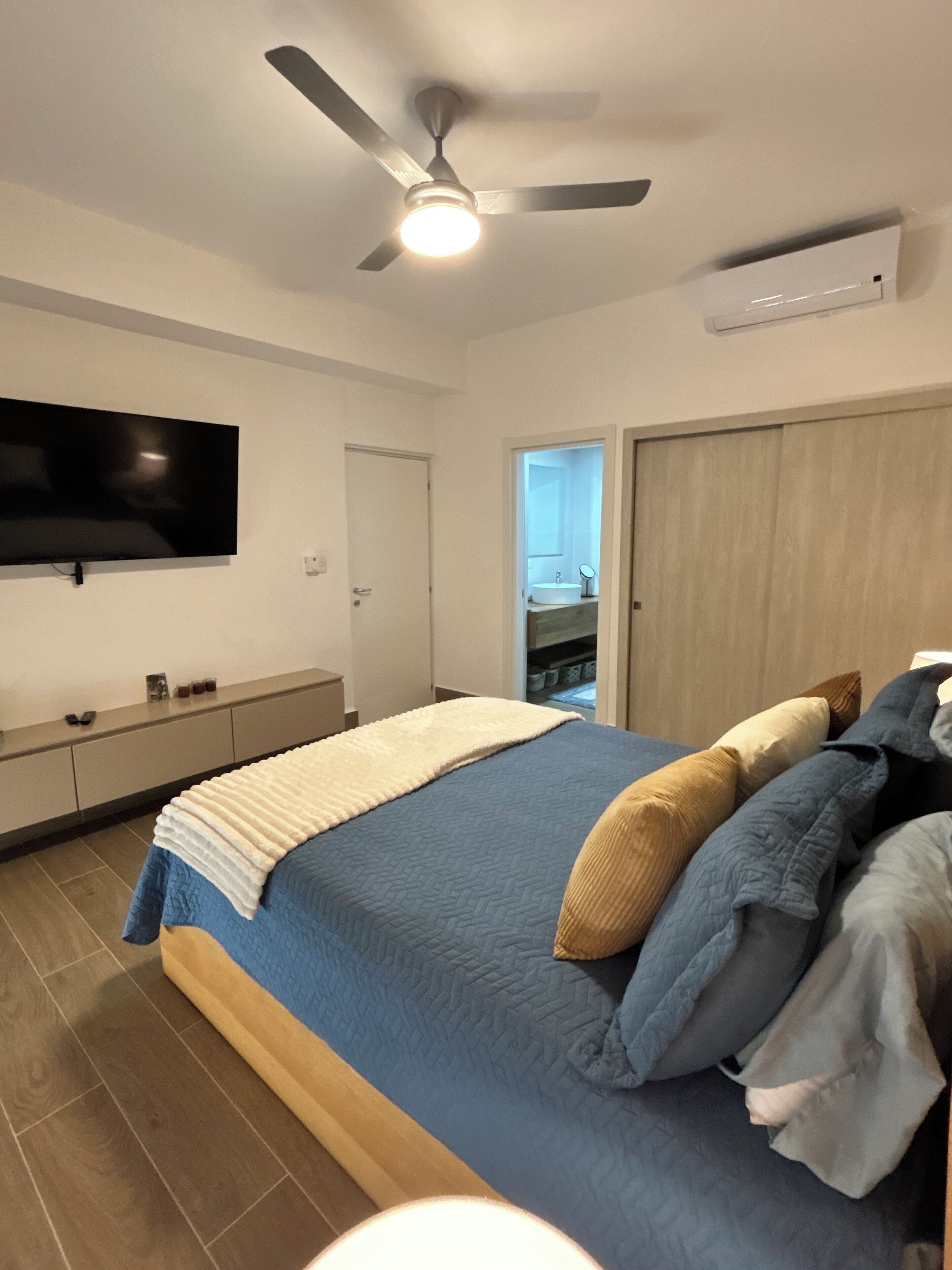 Ein Schlafzimmer mit einem Bett, einem Deckenventilator, einem an der Wand montierten Fernseher, einem Kleiderschrank und einem durch eine offene Tür einsehbaren Badezimmer.