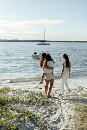 A beach scene with a boat on the water and people standing on the shore. - Stone & Tide by Aryve (Runaway Bay)