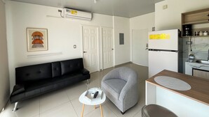 Ein gemütlicher Wohnbereich mit einem schwarzen Sofa, einem grauen Sessel, einem kleinen weißen Tisch und einer Küchenzeile.