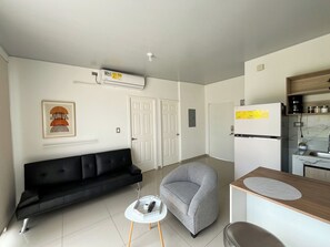 Ein gemütlicher Wohnbereich mit einem schwarzen Sofa, einem grauen Sessel, einem kleinen weißen Tisch und einer Küchenzeile.