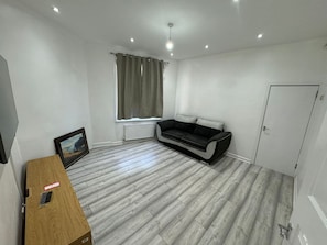 Ein Zimmer mit einer grauen Couch, einem Holztisch, einem Fernseher und einem Fenster mit Vorhängen.