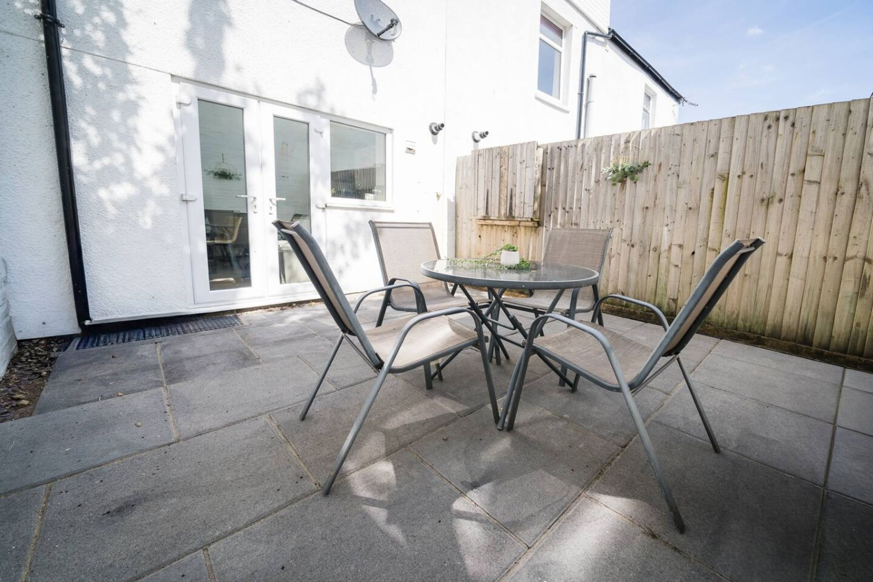 A patio area with a round table and four chairs, a wooden fence, and a white building with a satellite dish.
