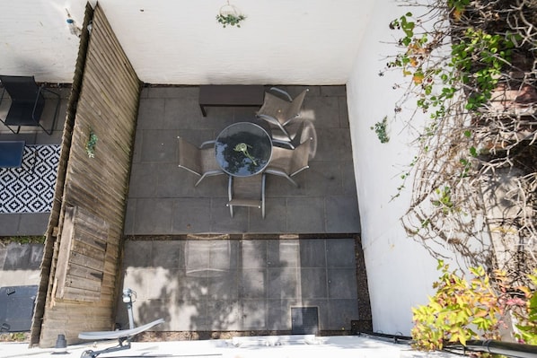 Uma área de estar moderna ao ar livre, com mesa de vidro, cadeiras de metal, cerca de madeira e piso estampado.