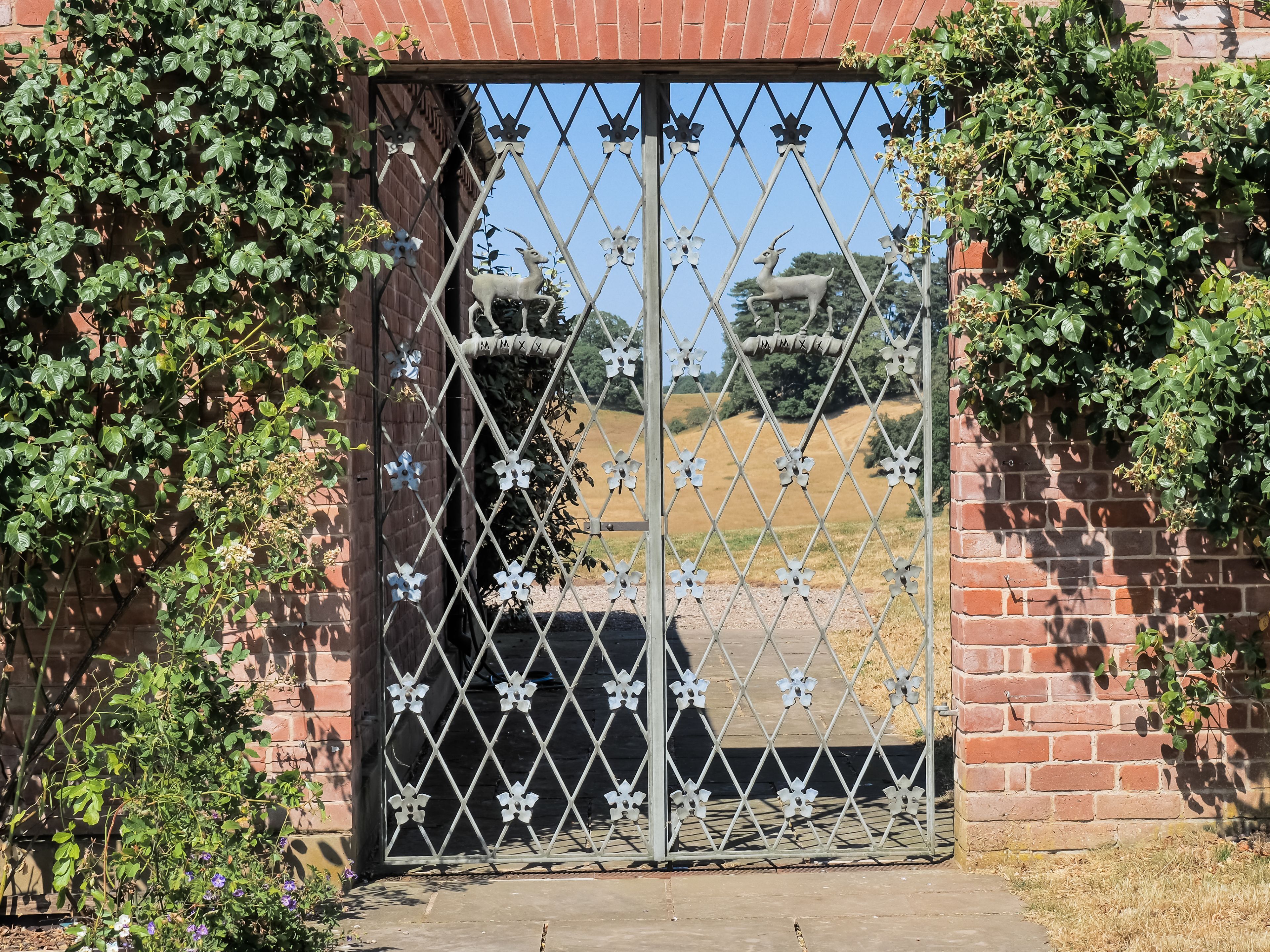 Une porte en métal ornée d’un motif décoratif, un mur de briques et de la verdure de chaque côté.