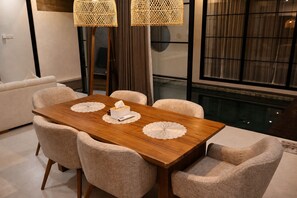 Uma área de refeições com mesa de madeira, quatro cadeiras estofadas e duas luminárias pendentes de fibra.