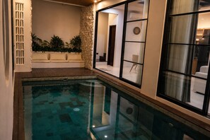 A swimming pool with a wooden deck, a stone wall with plants, and a view of a room with a clock and a chair. - Cala Vista Villa Tumbak Bayuh Bali. Spacious 3BR Villa for Families & Groups. (Tumbak Bayuh)