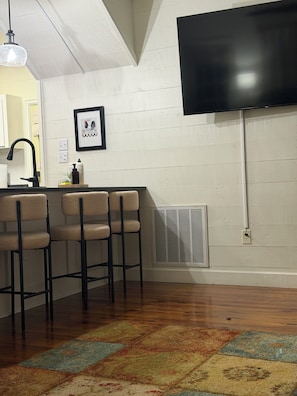 A kitchen with a bar area, a large flat-screen TV mounted on the wall, and a patterned rug on the floor. - Over the Creek and Thru the Woods. Comfy 2BR/2bath. Sleeps9 .Firepit/pond (Abbeville)