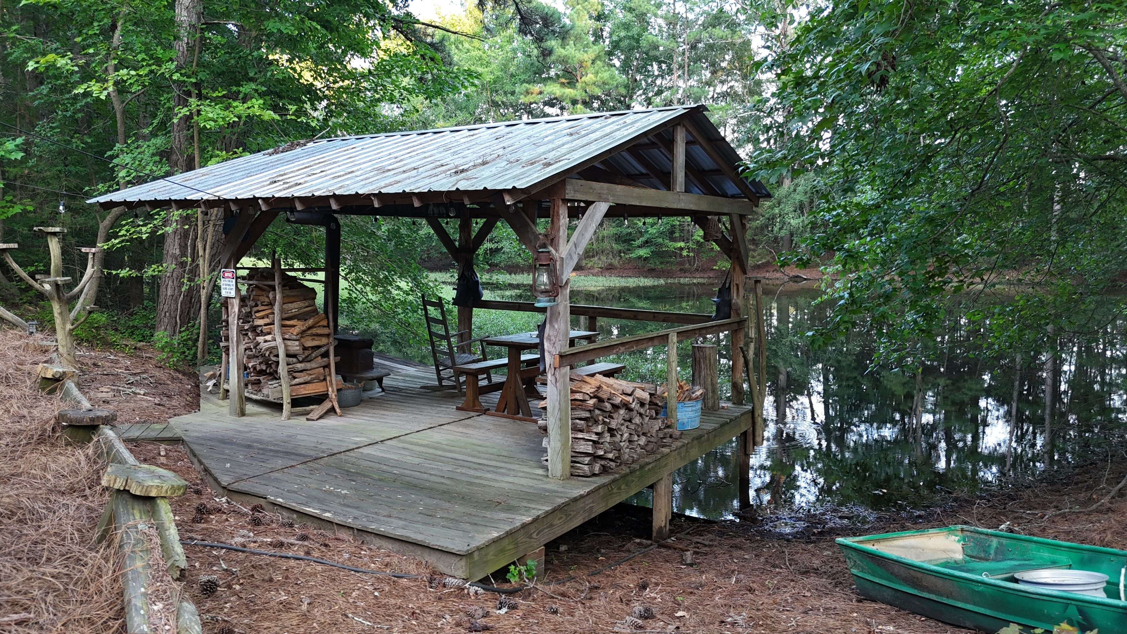 Eine Holzterrasse mit Metallüberdachung, umgeben von Bäumen und einem Gewässer.