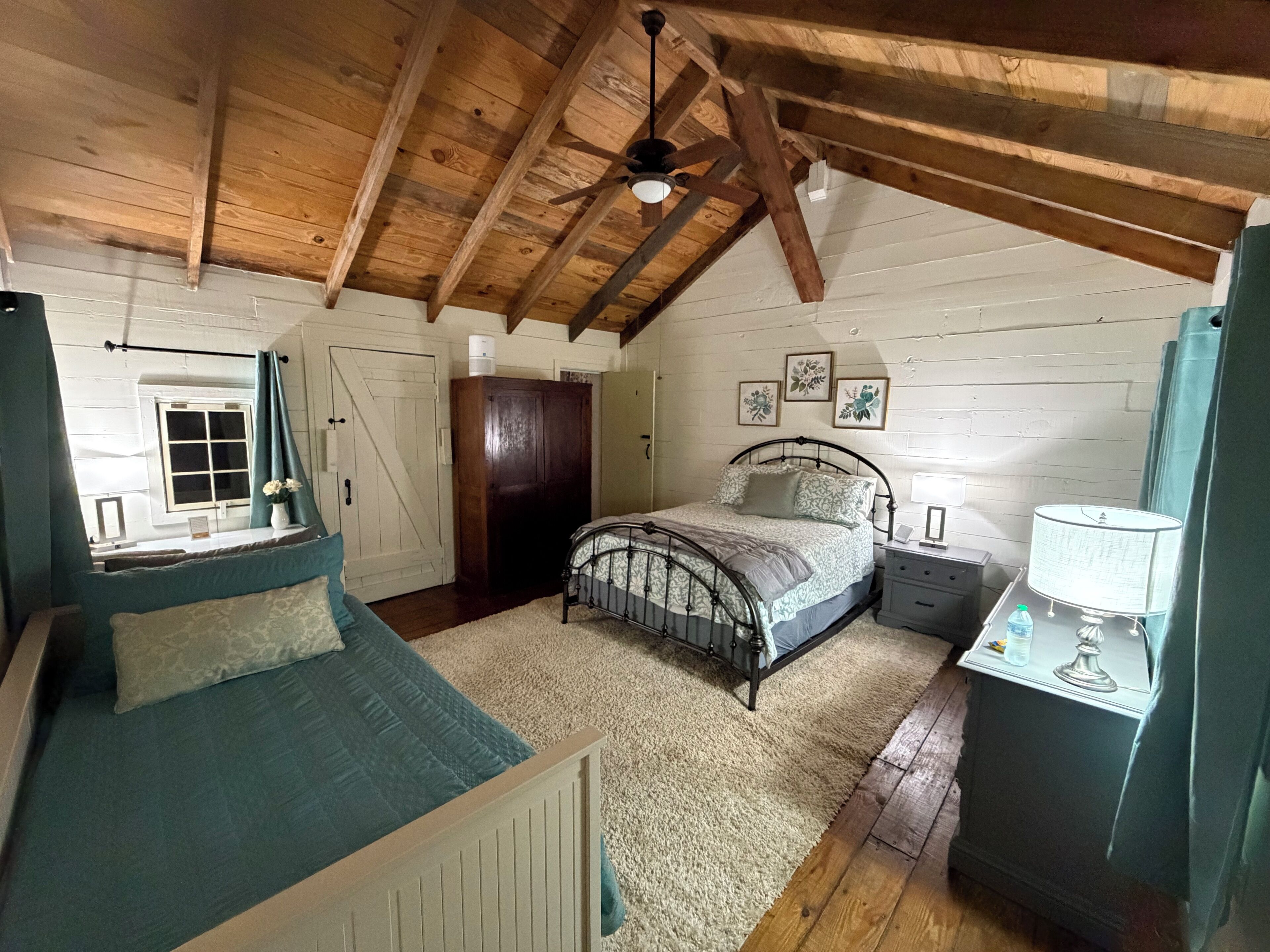 Een slaapkamer met een schuine plafond, een bed, een nachtkastje, een bureau met een lamp en een raam met gordijnen.